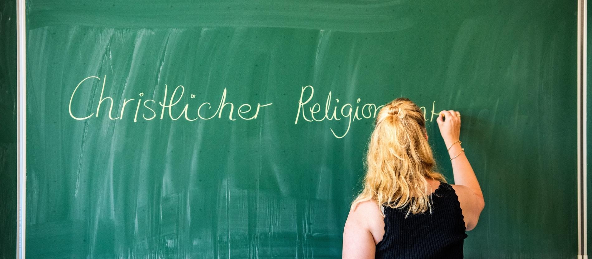 Eine Lehrerin schreibt den Schriftzug "Christlicher Religionsunterricht" mit Kreide an eine Tafel.