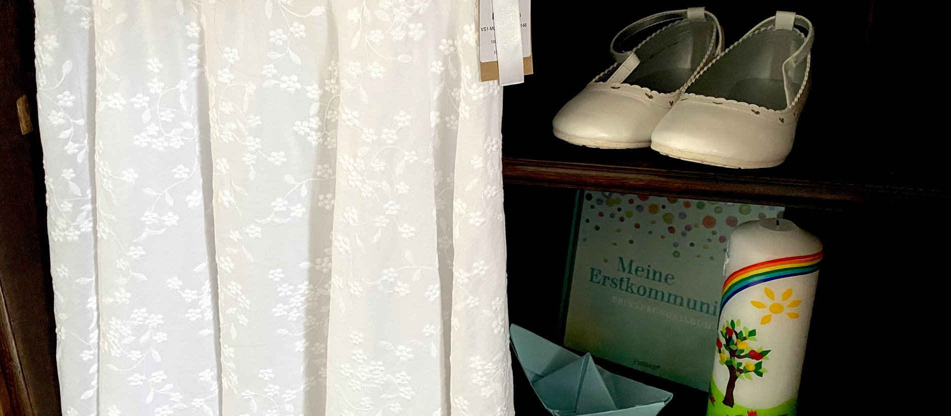 Ein weißes Kommunionkleid hängt vor einem Regal, in dem Kommunionschuhe, eine Taufkerze, ein Papierschiffchen und ein Buch über die Erstkommunion stehen.