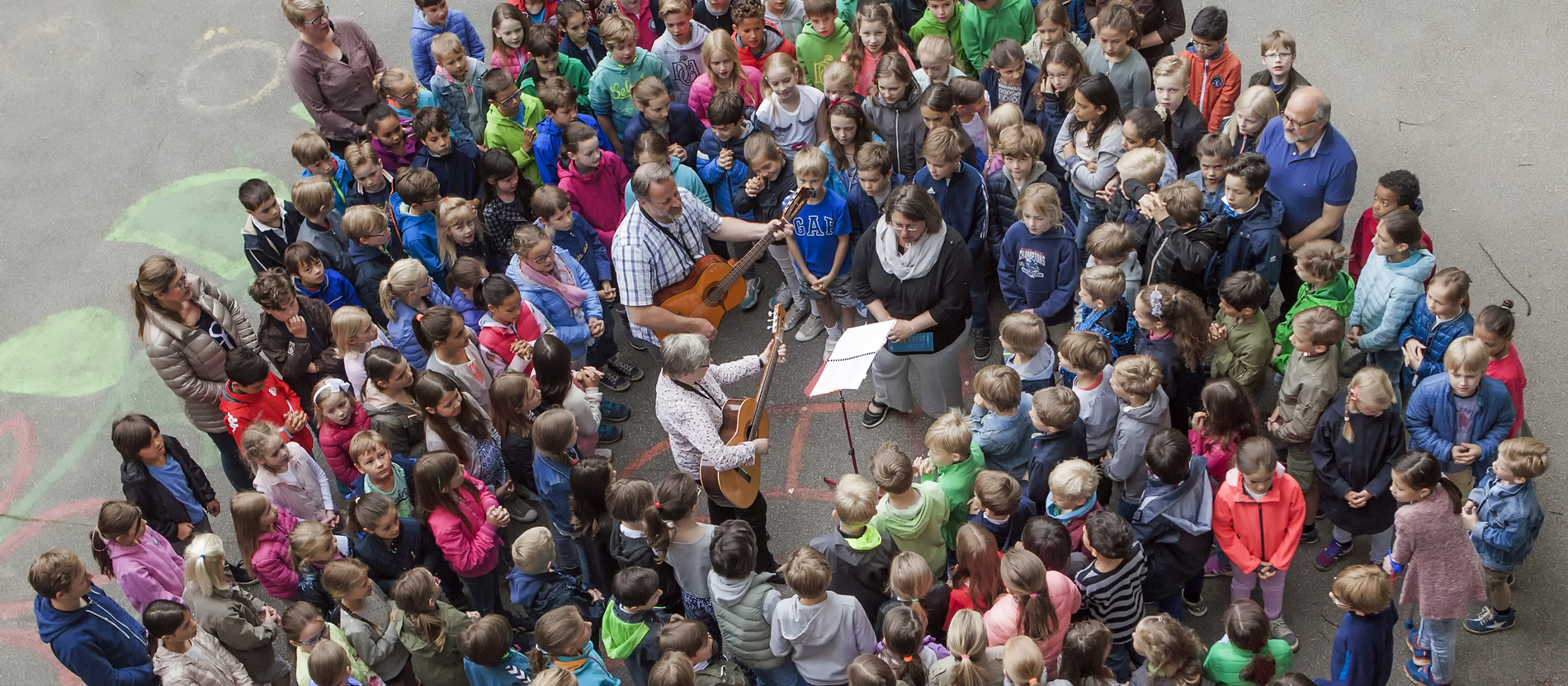 Hunderte Grundschulkinder stehen auf dem Schulhof rund um Lehrer/innen, die mit Gitarre Lieder singen und beten.