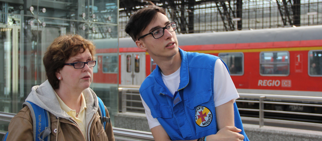 Eine Frau in brauner Jacke steht neben einem Mitarbeiter der Bahnhofsmission, der auf eine weit entfernte Videotafel (nicht im Bild) schaut.