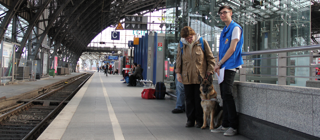 Der Kölner Hauptbahnhof. Am rechten Bildrand stehen eine Frau in einer braunen Jacke und ein junger Mann in einer Weste der Bahnhofsmission. Zwischen den beiden sitzt ein Schäferhund.