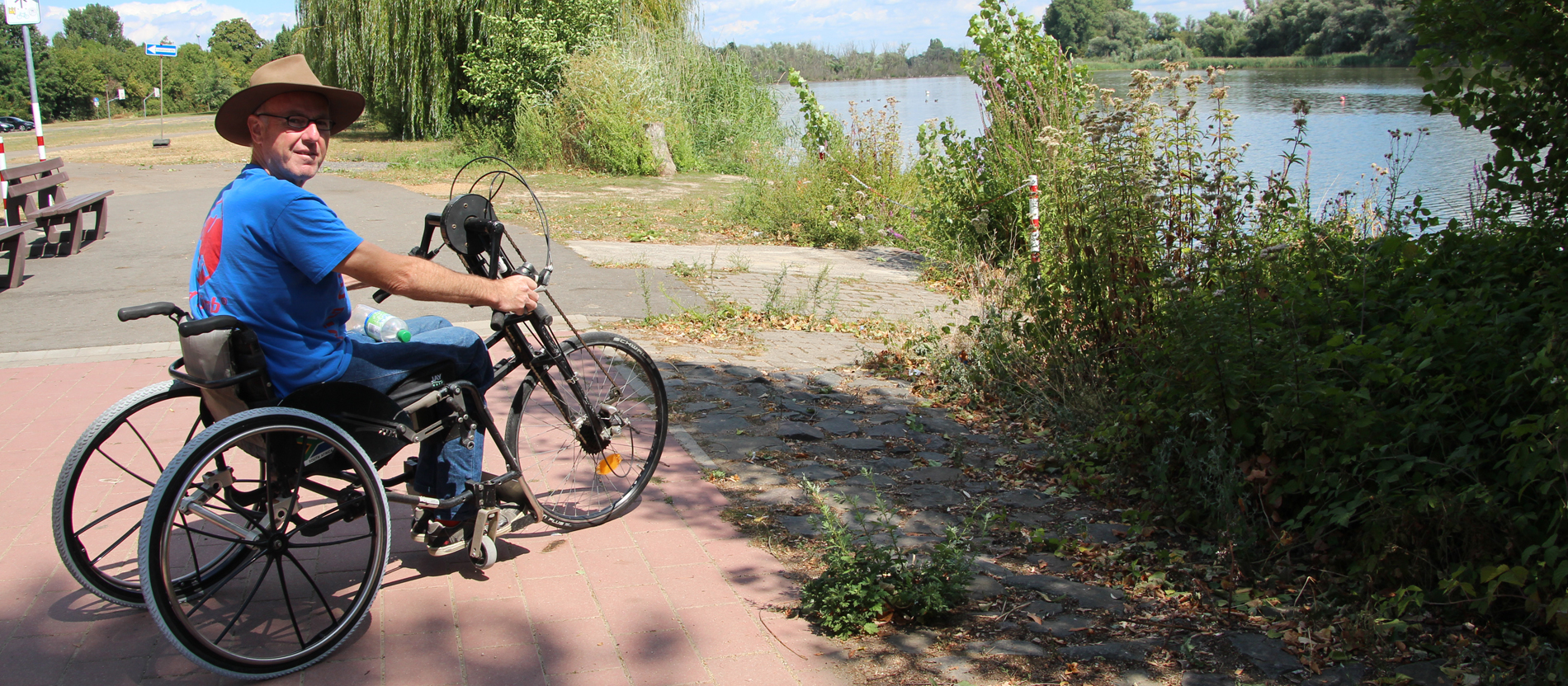 Ein Rollstuhlfahrer steht mit seinem Handbike an einem See