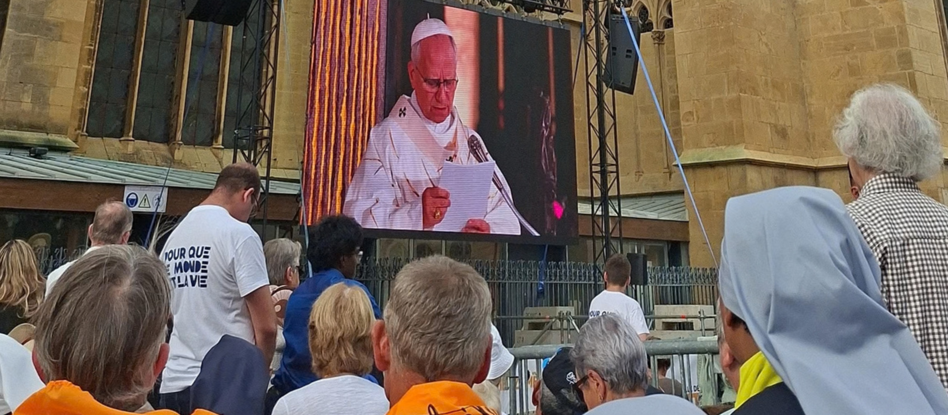 Auf einem großen Bildschirm ist Papst Leo XIV. zu sehen. Er predigt mit einem Zettel in der Hand. Vor dem Bildschirm stehen viele Menschen.