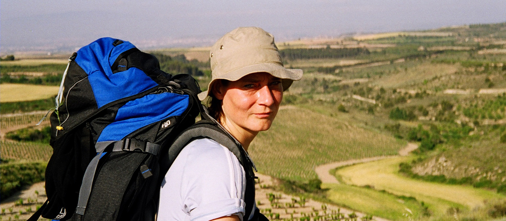 Frau mit Hut und Rucksack. Im Hintergrund weite Landschaft.