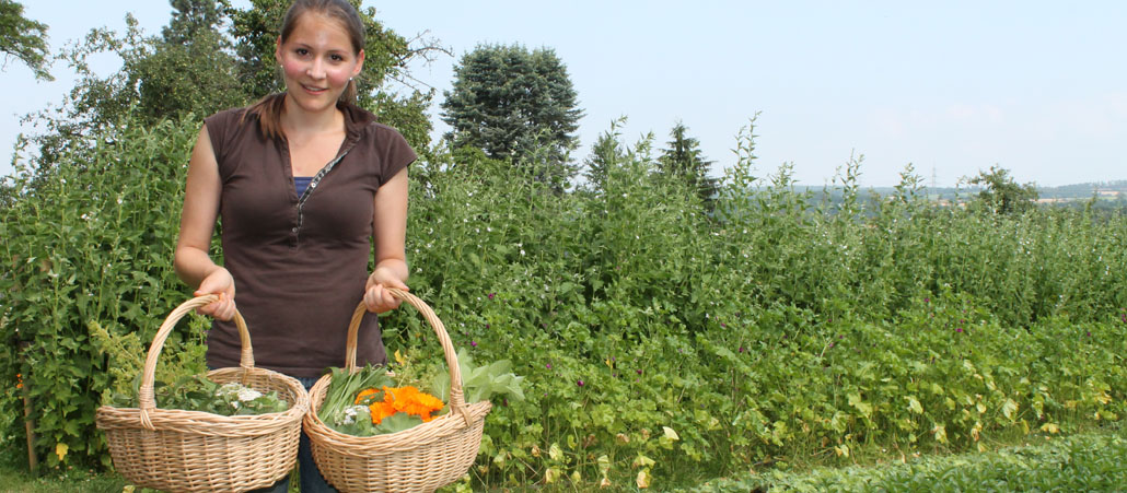 Eine Frau steht mit zwei Körben mit Kräutern im einem großen grünen Garten.
