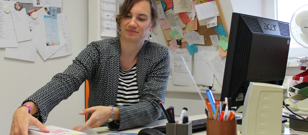 Eine Frau sitzt in einem Büro an einem Schreibtisch, vor ihr Unterlagen und ein PC.