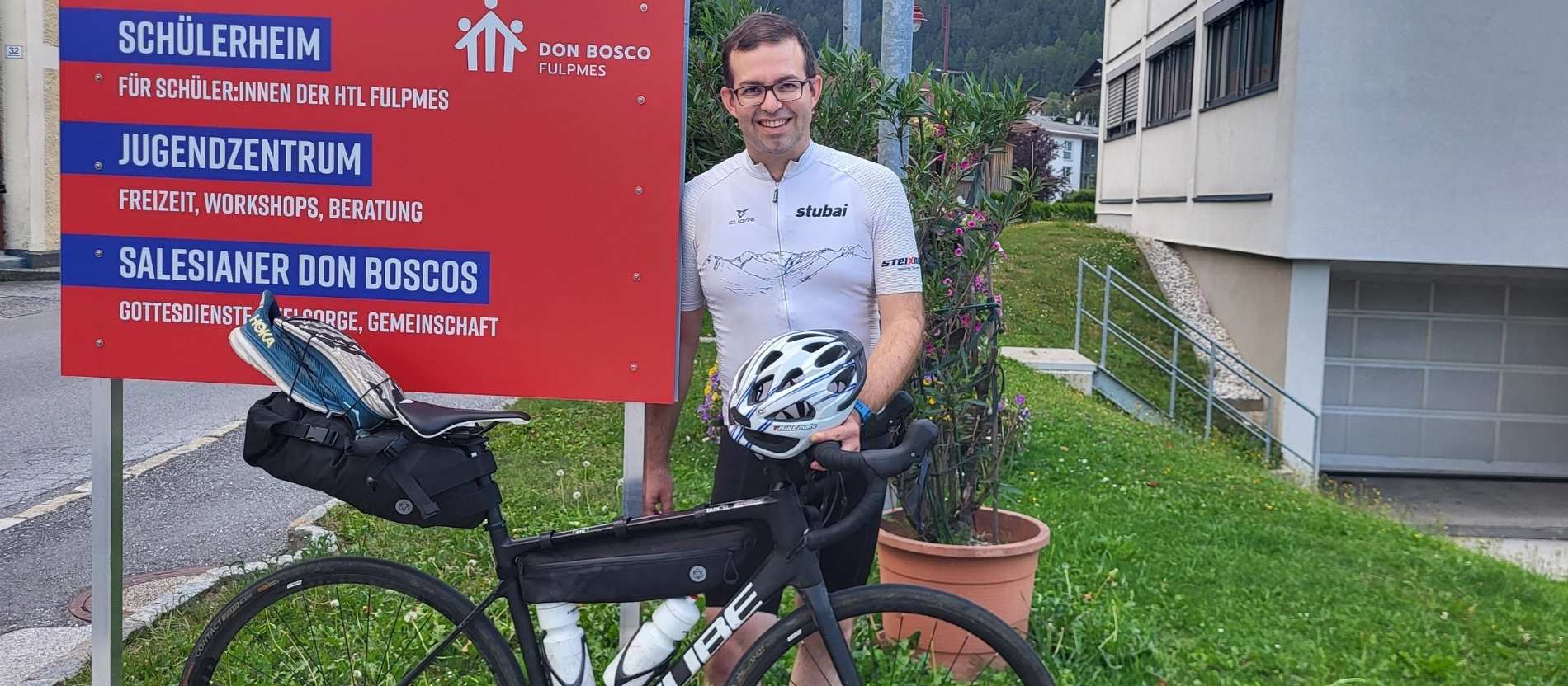 Pater Peter Rinderer mit einem Fahrrad neben einem Schild einer Don-Bosco-Einrichtung in Fulpmes