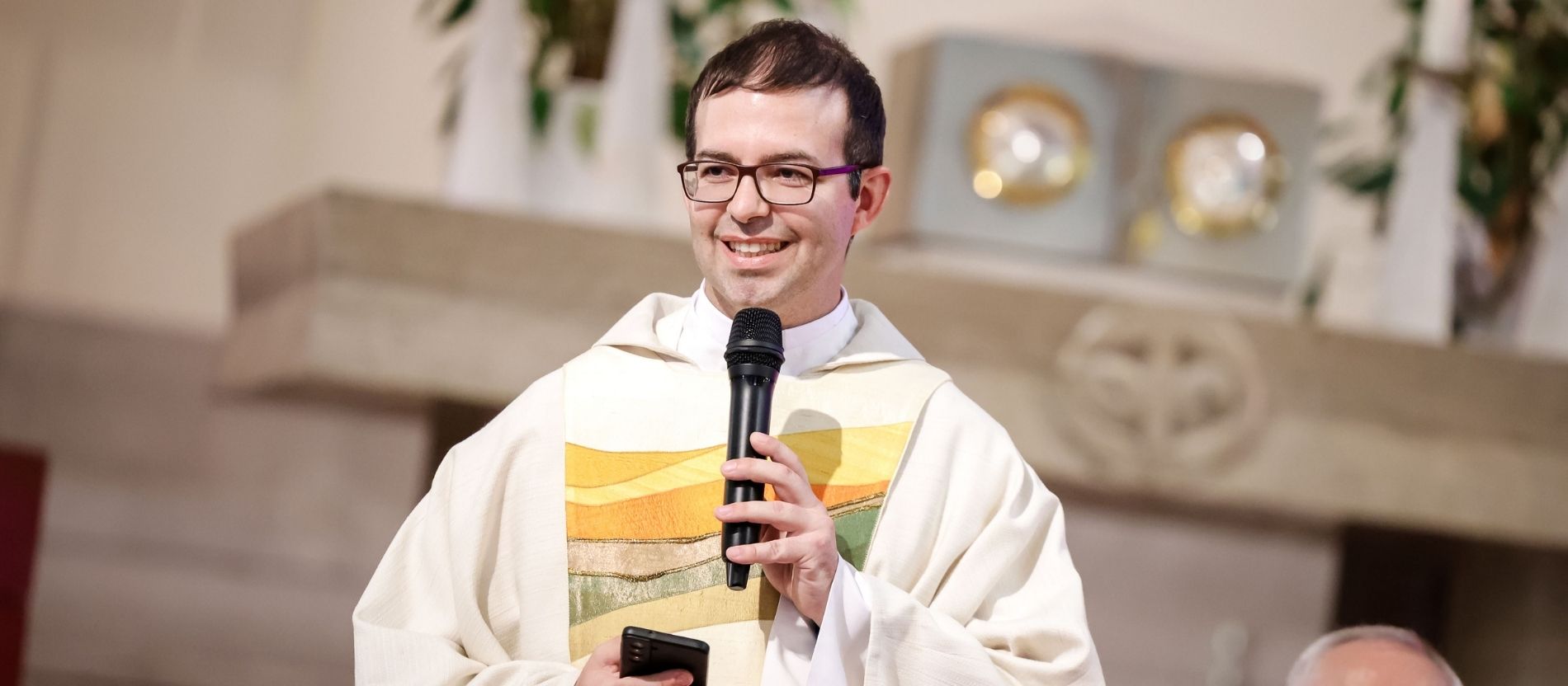 Pater Peter Rinderer spricht bei einem Gottesdienst in ein Mikrofon, in der Hand hält er ein Smartphone
