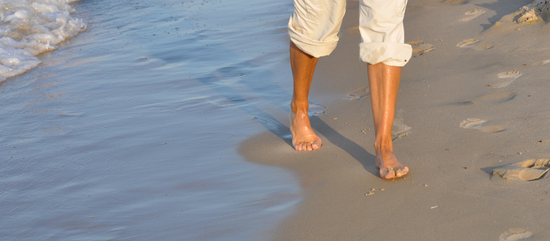 Eine Frau spaziert barfuß am Strand entllang.