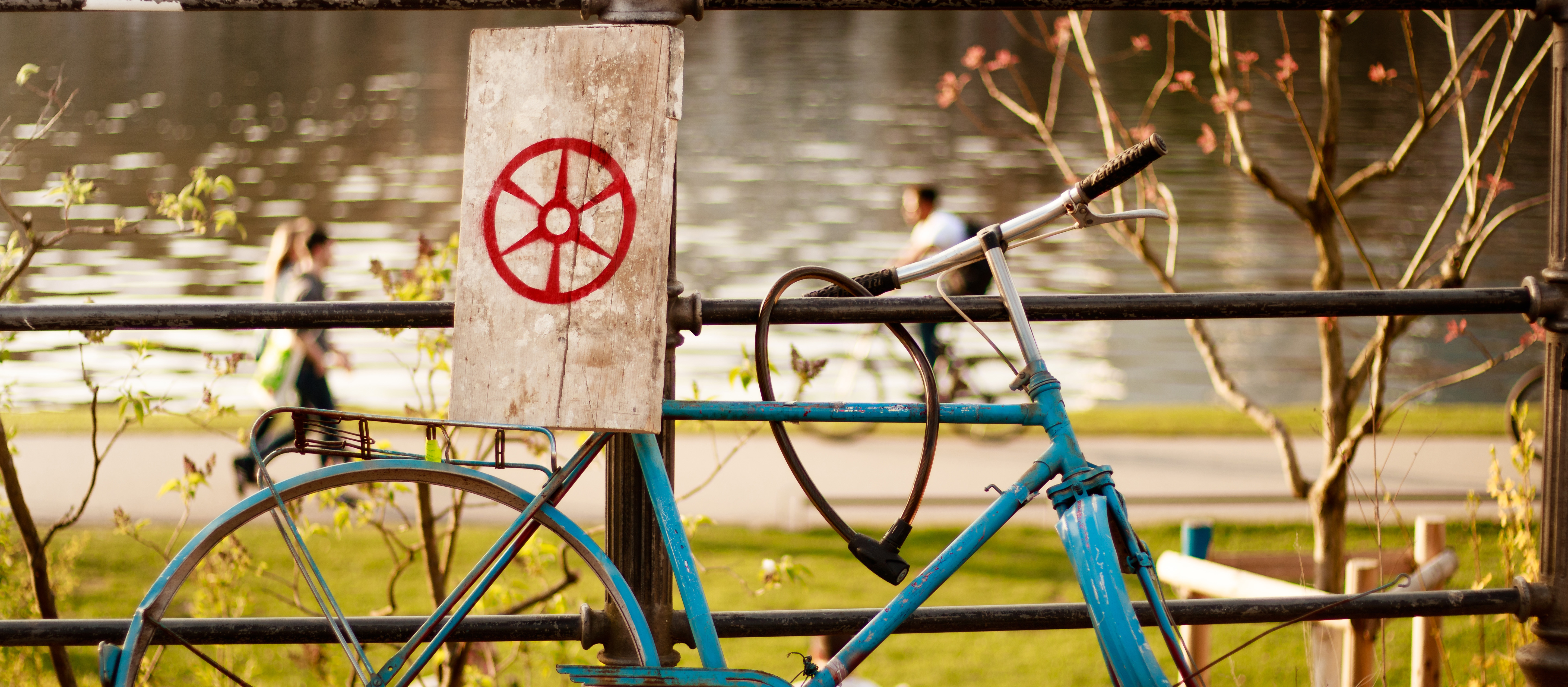 Ein blaues Fahrrad ist an ein Geländer angelehnt, am Sattel ist ein Holzbrett befestigt, auf dem ein rotes Steuerrad abgebildet ist. Im Hintergrund ist ein Flussufer zu sehen. 