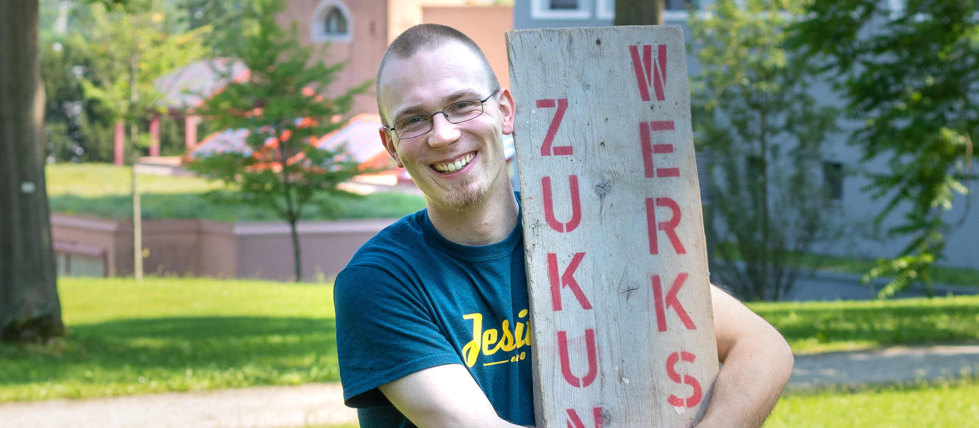 Der Jesuit Clemens Kascholke hält lachend ein Holzbrett in den Armen, auf dem in roten Buchstaben Zukunftswerkstatt steht 